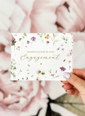 A beautifully designed engagement card with delicate floral watercolor illustrations, featuring the message "Congratulations on Your Engagement" in elegant script. The card is styled on a soft white fabric background with a sprig of greenery beside it.